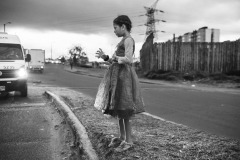 A young girl looks at the empty plastic glass she recollects the alms in, along a street of the capital city Bogotà, on October 31, 2018. 
In October and November 2018, the photographer documented the living conditions of Venezuelan refugees and migrants in an area close to the capital city's bus terminal, where approximately three hundred people were living in makeshift tents.
On the day of this photograph, compassionate Colombians had come to the settlement to cheer the children up by distributing Halloween dresses. A young girl was begging for money and exchanging candies for coins with the drivers passing by the street. Like other children, she ran to receive a dress, quickly wore it, and walked toward the traffic island again. 
In November, Colombian authorities dismantled the camp, forcing people to continue their journey to other Colombian cities or the southern border between Colombia and Ecuador. Others resettled in an official humanitarian settlement, also dismantled a month later. The photographer was, by the time, well known by the families in the camp, who granted their consent to work.